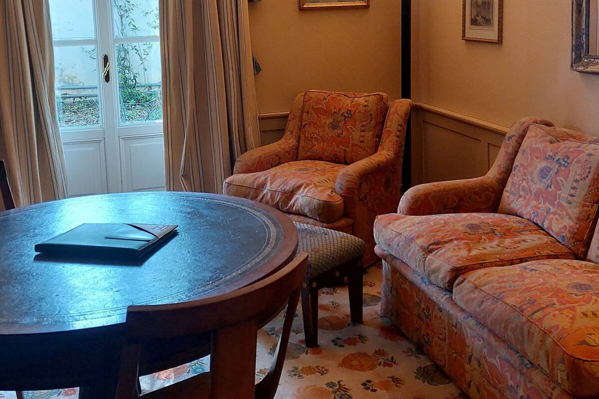 A cozy living room with a round wooden table, two patterned armchairs, a sofa, and a door leading to a balcony.