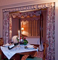 Una stanza con un letto a baldacchino, un tavolo apparecchiato per due con vino e bicchieri, e una candela accesa.