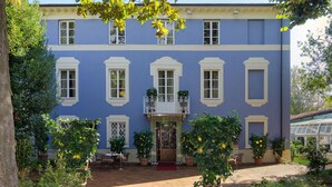 A two-story building with a blue facade, white window frames, and a central entrance with a red carpet. There are potted plants with yellow flowers in front of the entrance. - Resort Dei Limoni (Lucca)