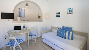 A bedroom with a bed, a desk with a laptop, a chair, a TV, and a ceiling fan. - Gryparis Club Apartments (Mykonos)