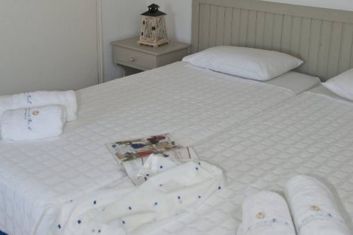 A neatly made bed with white linens, a small table with a magazine, and a nightstand with a lamp.