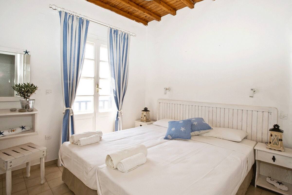 A bedroom with two beds, a wooden ceiling, a window with curtains, a bedside table with a lamp, and a small shelf with decorative items.