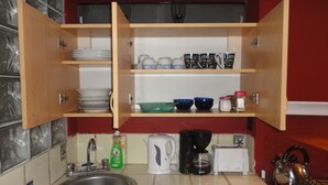Una cocina con lavabo, cafetera, hervidor y armarios abiertos con vajilla y tazas.