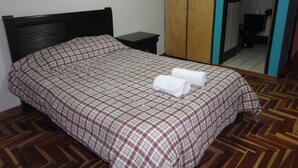 Una cama bien hecha con una colcha a cuadros y dos toallas enrolladas encima, un armario de madera, una mesita de noche pequeña y un espejo en la pared.