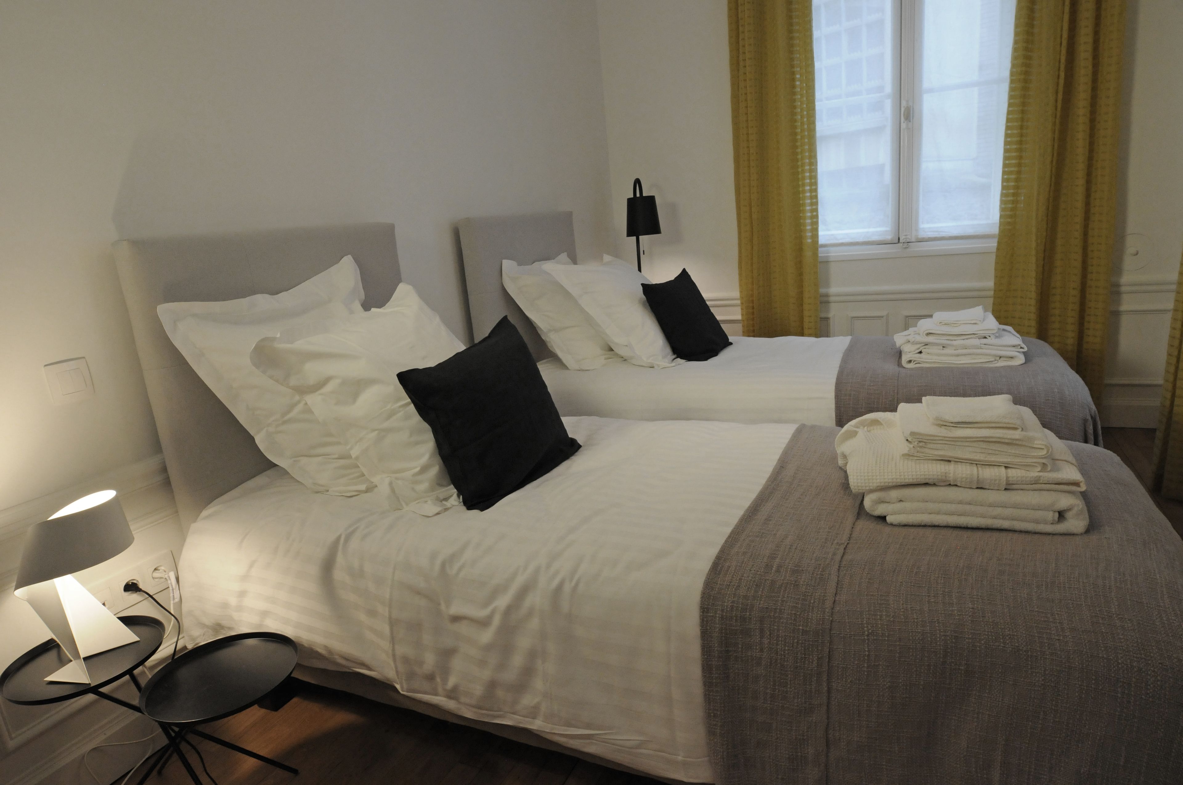 A hotel room with two beds, a bedside table with a lamp, and a window with curtains.