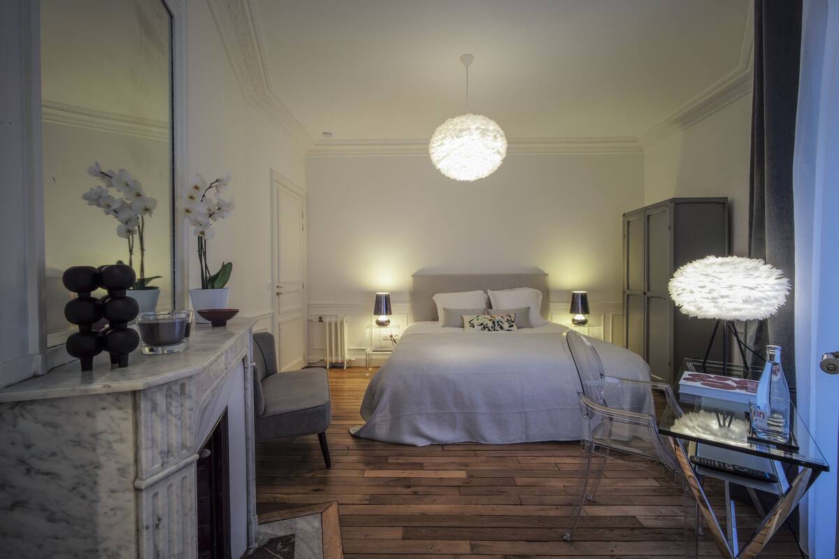 A modern bedroom with a large bed, a sitting area with a chair, a glass table, a fireplace, and a mirror.