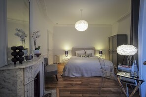 Een moderne slaapkamer met een groot bed, een zithoek met een fauteuil, een glazen tafel, een open haard en een spiegel.