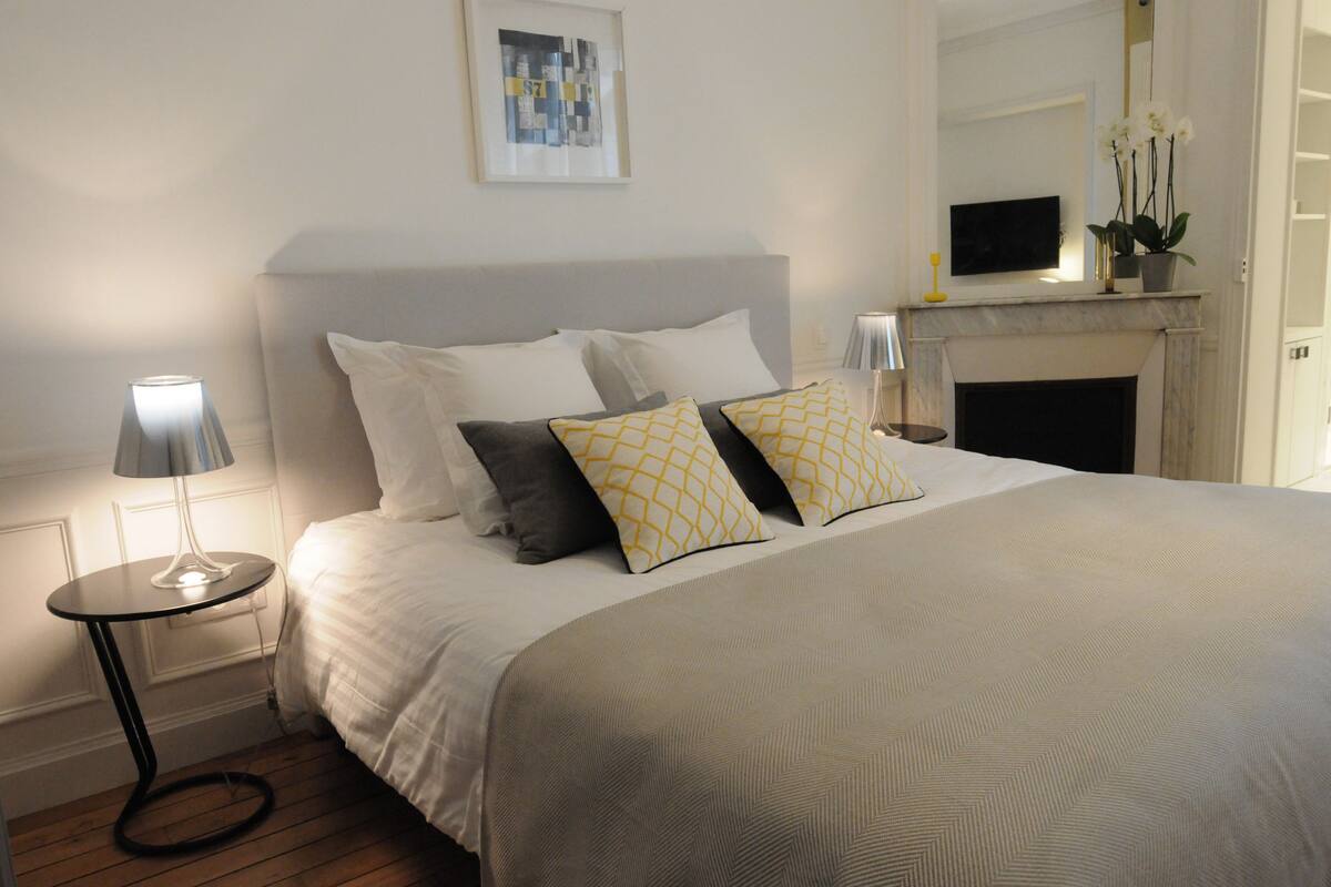 A neatly made bed with a grey headboard, a bedside table with a lamp, and a fireplace with a vase of flowers.