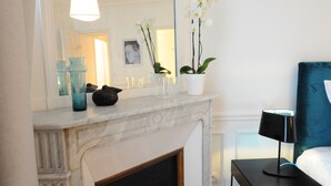 A room with a marble fireplace, a bed with a blue headboard, a nightstand with a lamp, and a mirror reflecting the room. - Relais 12bis B&B (Paris)