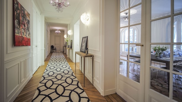 A hallway with a patterned carpet, a chandelier, framed pictures on a console table, and a view of an outdoor dining area through large windows. - Relais 12bis B&B (Paris)