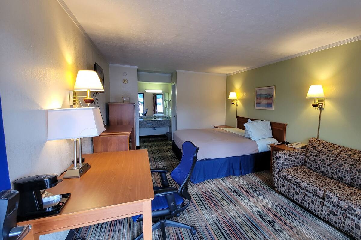 A hotel room with a bed, a sofa, a desk with a lamp, a chair, and a bathroom visible in the background.