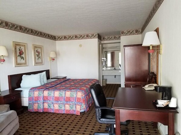 Habitación de hotel con cama, escritorio, silla y espejo. Hay dos lámparas, un teléfono y una cafetera en el escritorio.