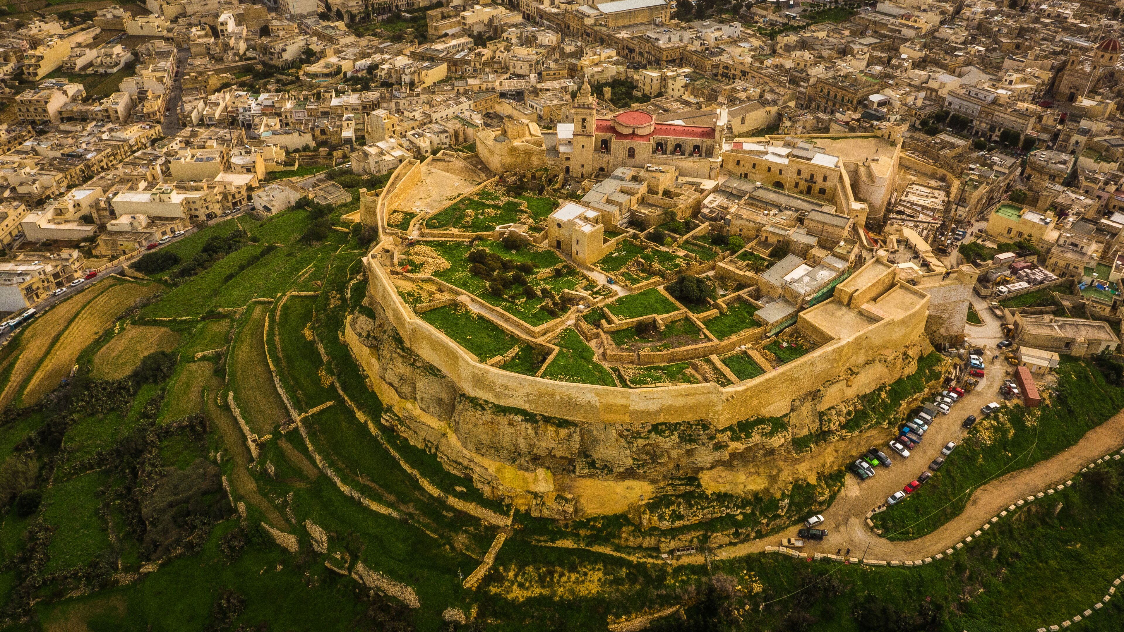 An aerial view of a historic walled city with a prominent fortress-like structure in the center, surrounded by residential buildings and narrow streets.