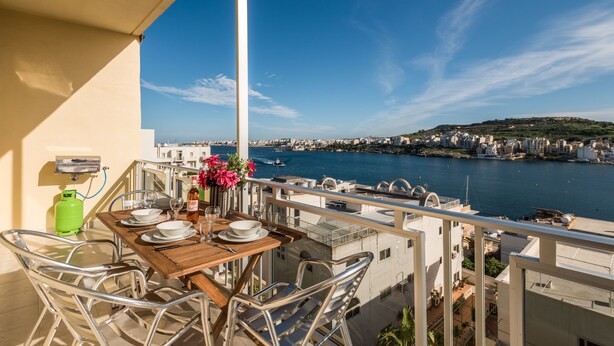 A balcony with a wooden table set for dining, overlooking a marina and cityscape. - Bay View Apartment by Getaways Malta (St. Paul's Bay)