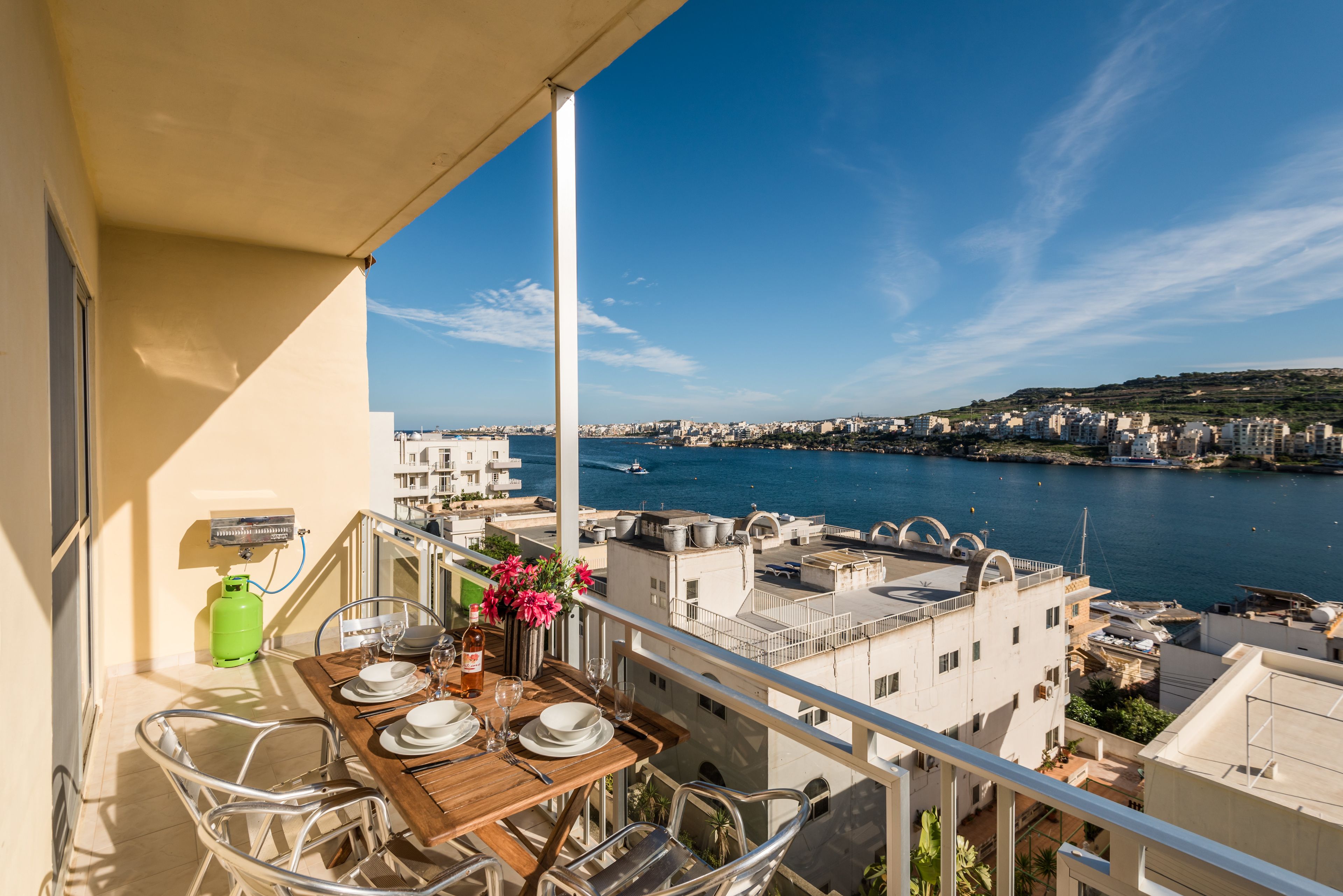 A balcony with a table set for dining, overlooking a coastal cityscape with buildings and a body of water.