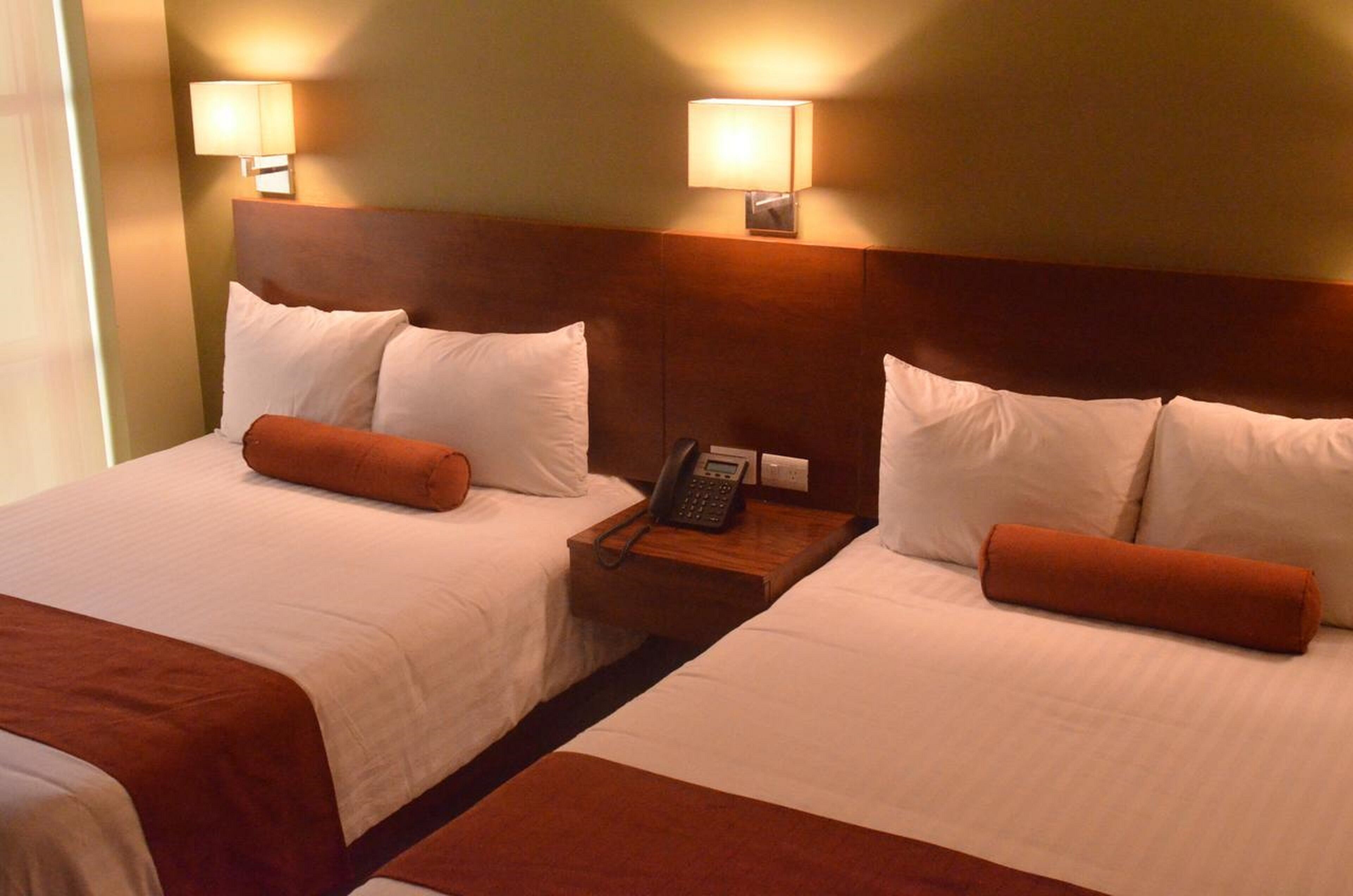 A hotel room with two beds, each with white linens and orange throw pillows. There are bedside lamps and a telephone on the nightstand between the beds.