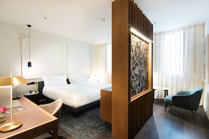 A hotel room with a large bed, a desk with a lamp, a chair, and a small table.
