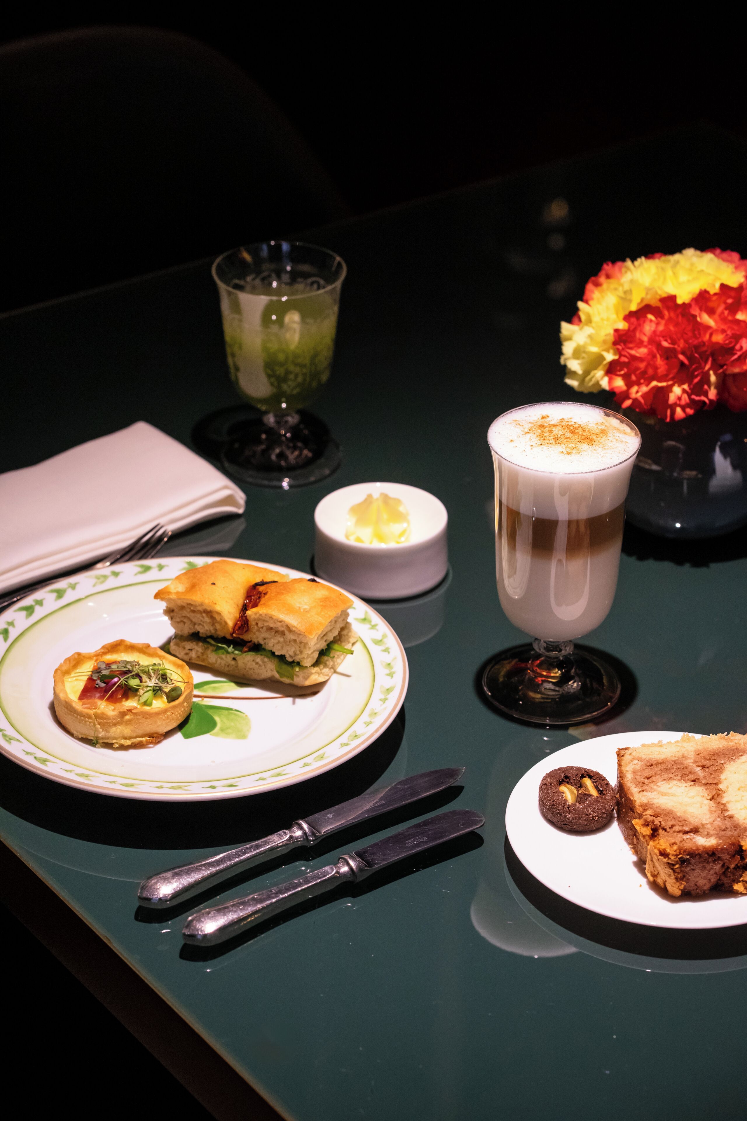 Una mesa con un sándwich, un vaso de bebida, un cuenco pequeño y un plato con un pastel.