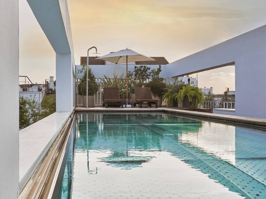 Una piscina con dos sillas de descanso y una sombrilla, rodeada de paredes blancas y plantas.