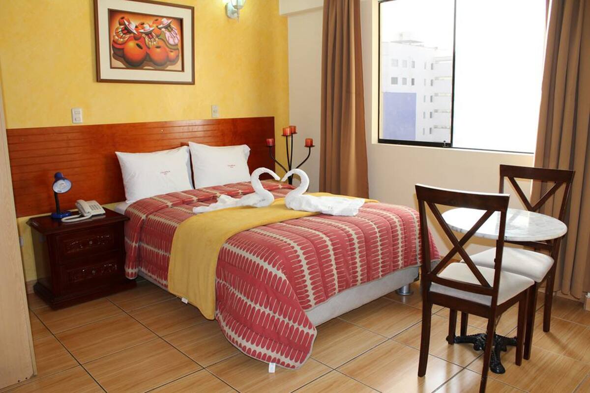 Habitación de hotel con cama, mesita de noche con lámpara, una silla, una pequeña mesa y ventana con vistas a edificios.