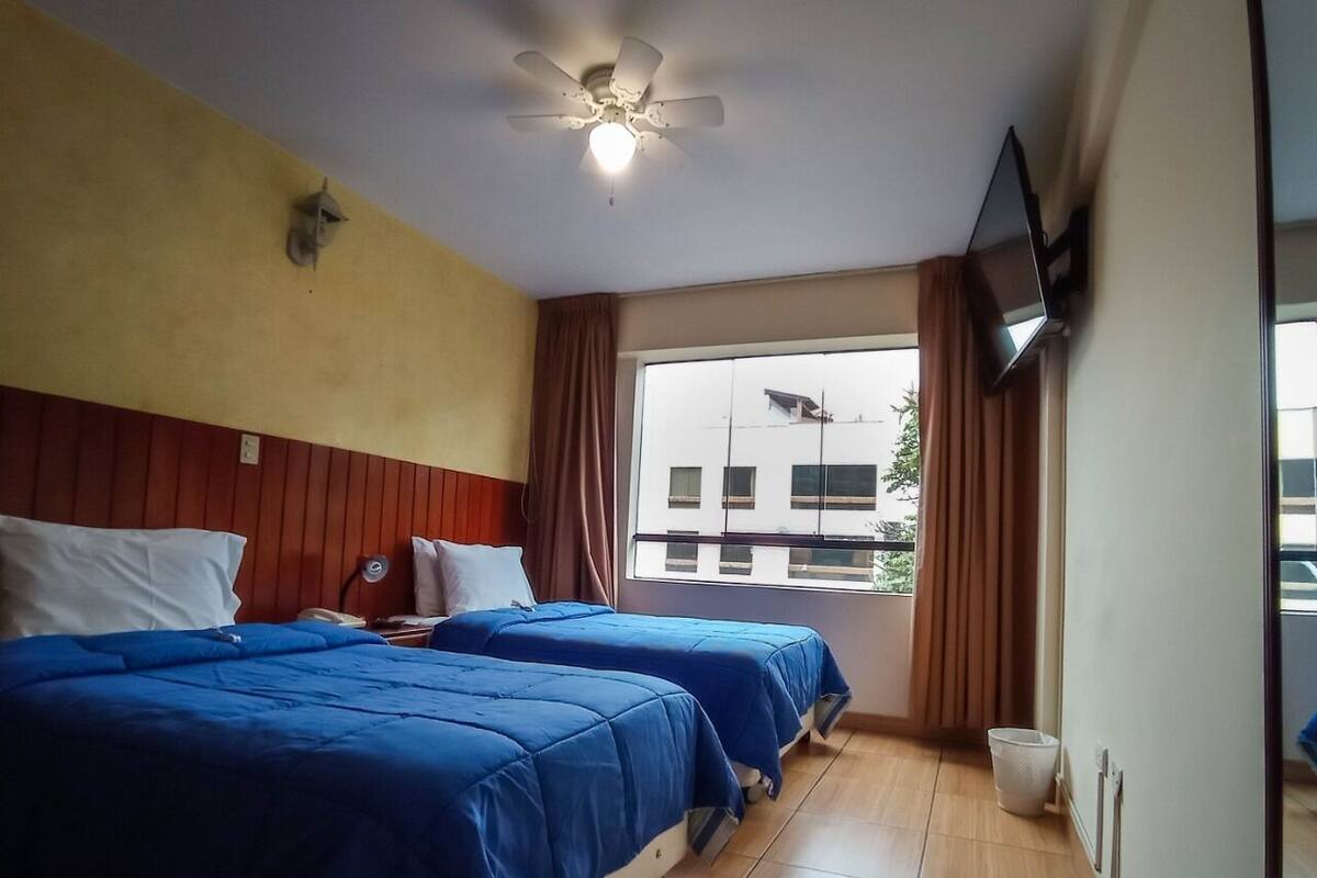 Habitación de hotel con dos camas, ventilador de techo, televisor montado en la pared y ventana con cortinas.