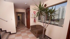 A hallway with a checkered floor, a brown sofa, a potted plant, and a window with a view of buildings. - Hotel El Farolito (Lima)