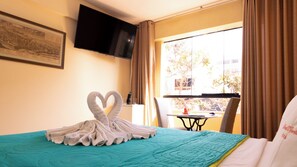 Habitación de hotel con cama, televisión de pantalla plana montada en la pared, ventana con cortinas y una mesa con una computadora portátil y una taza.