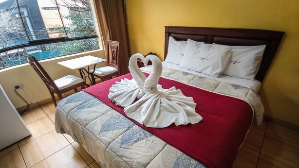 A hotel room with a bed, a red blanket, a swan towel art, a wooden chair, a small table, and a window with a view of buildings. - Hotel El Farolito (Lima)