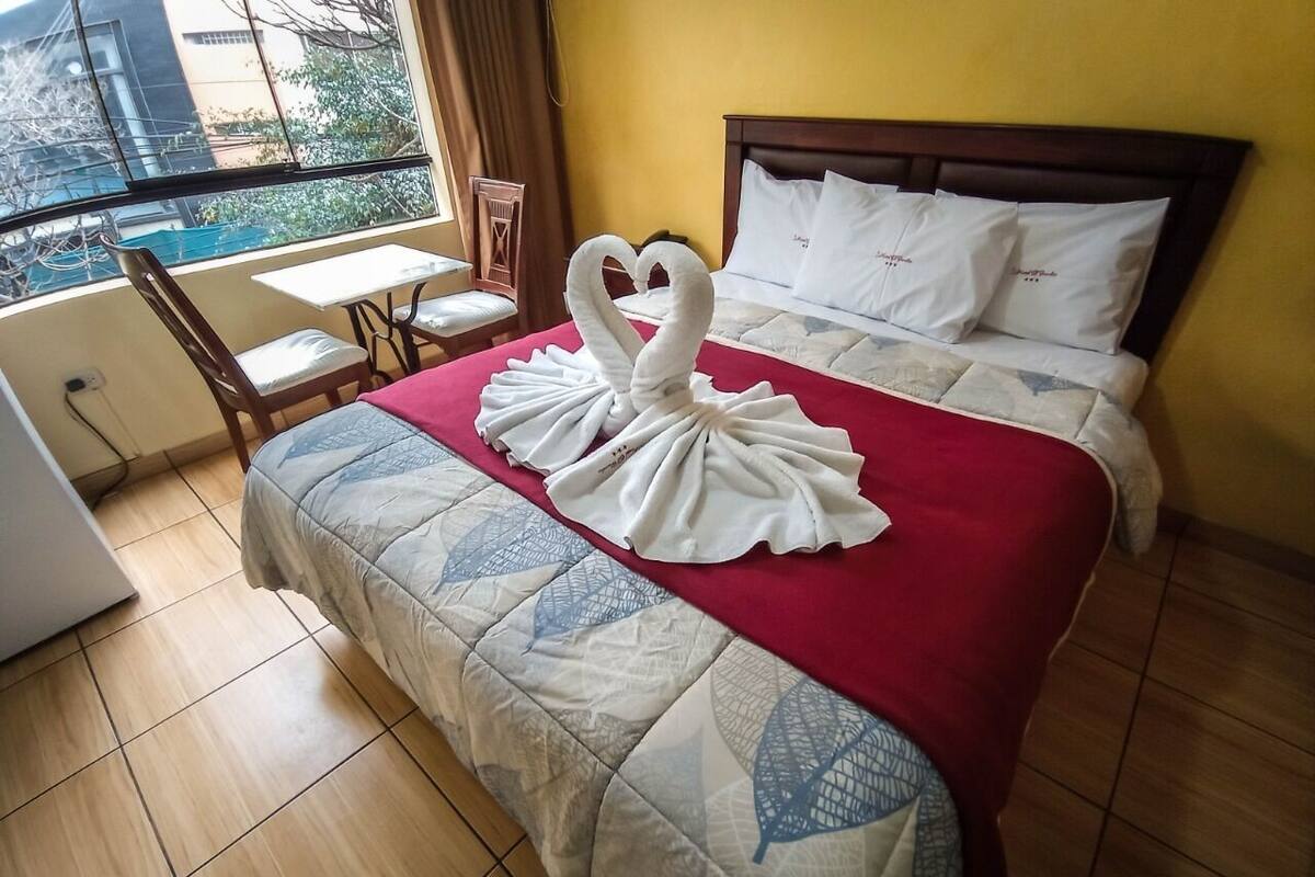 Habitación de hotel con cama, manta roja, figura de cisne hecha con toallas, una silla de madera, una mesita y ventana con vista a edificios.