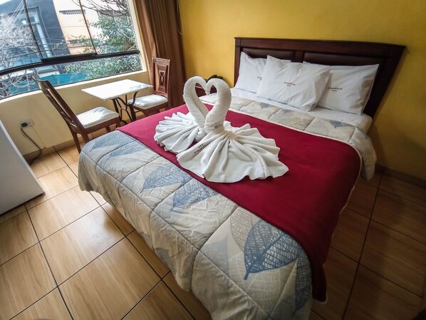 A hotel room with a bed, a red blanket, a swan towel art, a wooden chair, a small table, and a window with a view of buildings. - Hotel El Farolito (Lima)