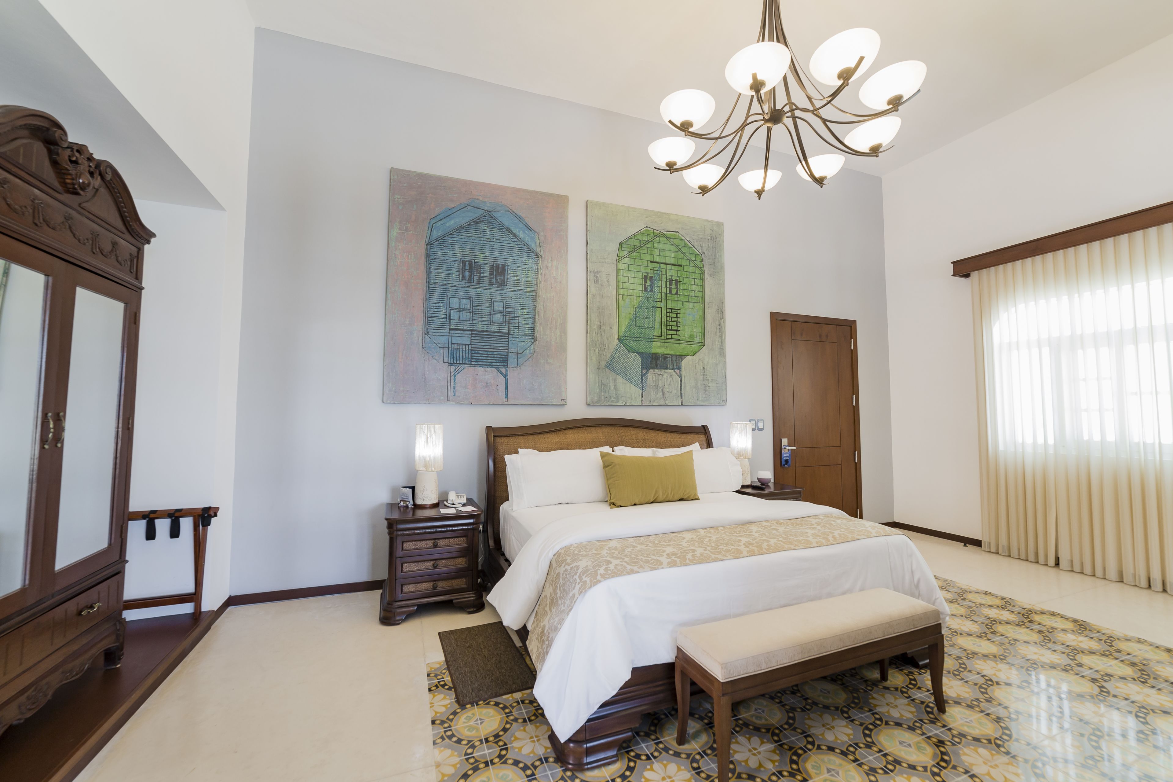 A bedroom with a large bed, wooden headboard, bedside tables with lamps, a bench, a chandelier, and two wall paintings.