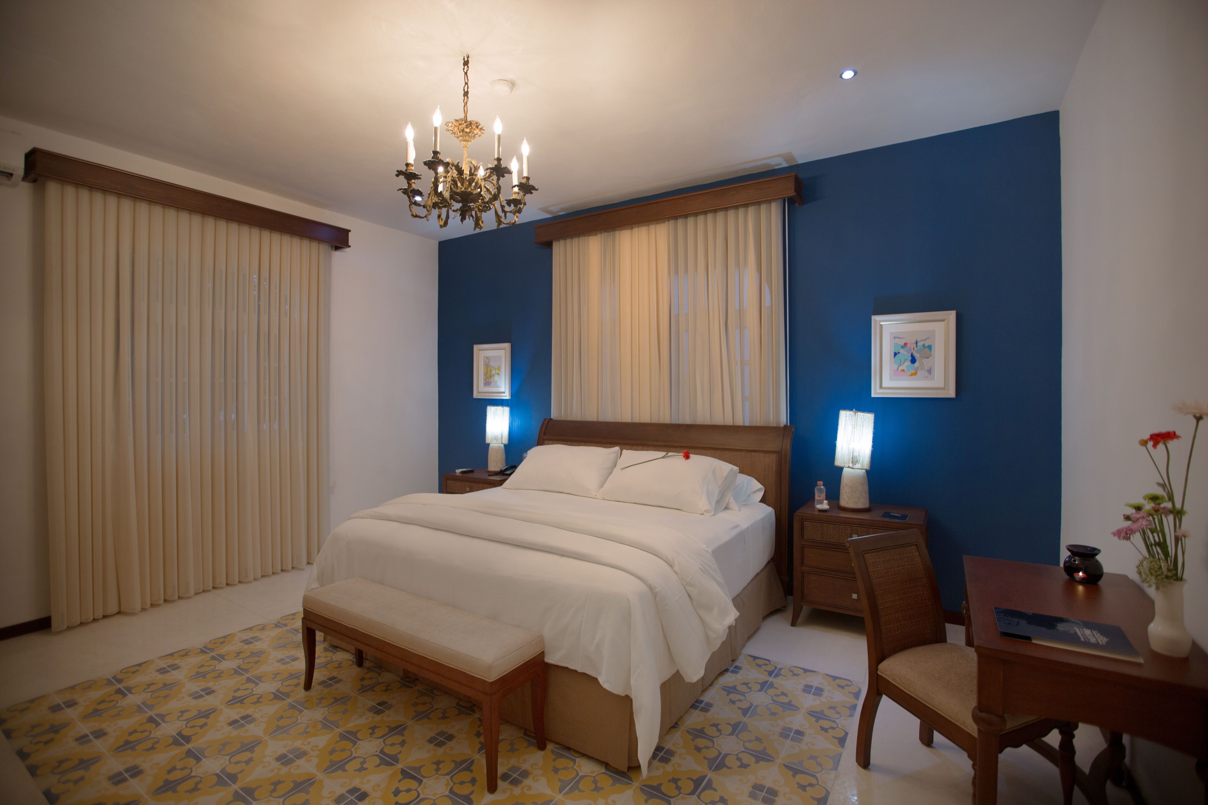 A bedroom with a large bed, wooden headboard, bedside tables with lamps, a desk with a chair, and a chandelier.