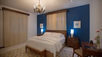 A bedroom with a large bed, wooden headboard, bedside tables with lamps, a desk with a chair, and a chandelier.