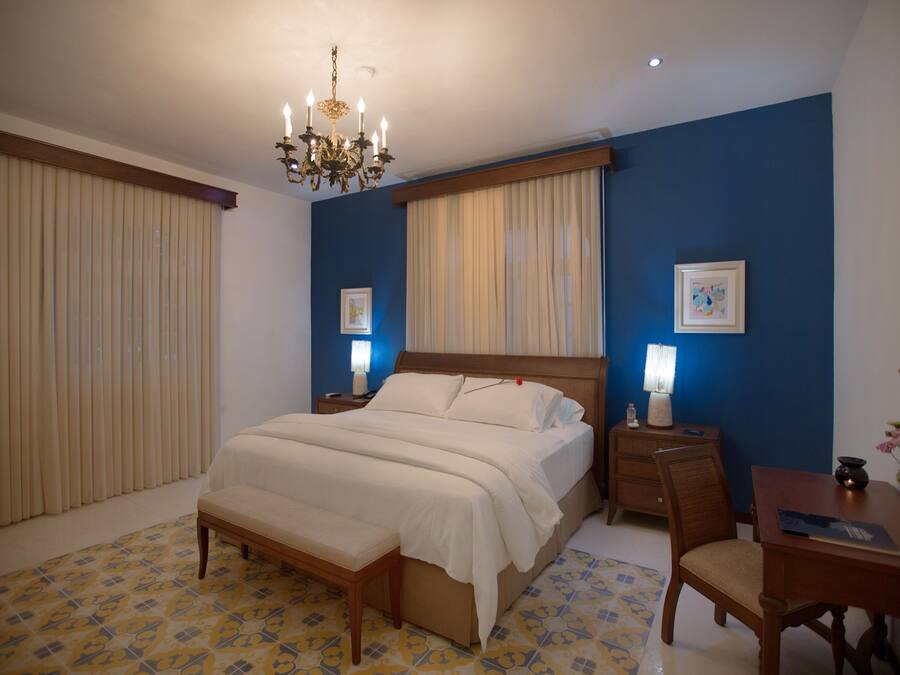 A bedroom with a large bed, wooden headboard, bedside tables with lamps, a desk with a chair, and a chandelier.