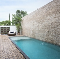A swimming pool with a stone wall, a tiled deck, and a seating area with cushions and plants.