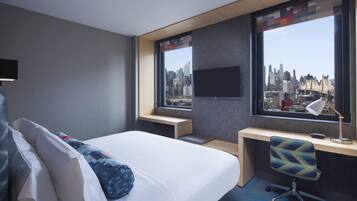 A hotel room with a bed, a desk, a chair, a TV, and a view of a city skyline through large windows.