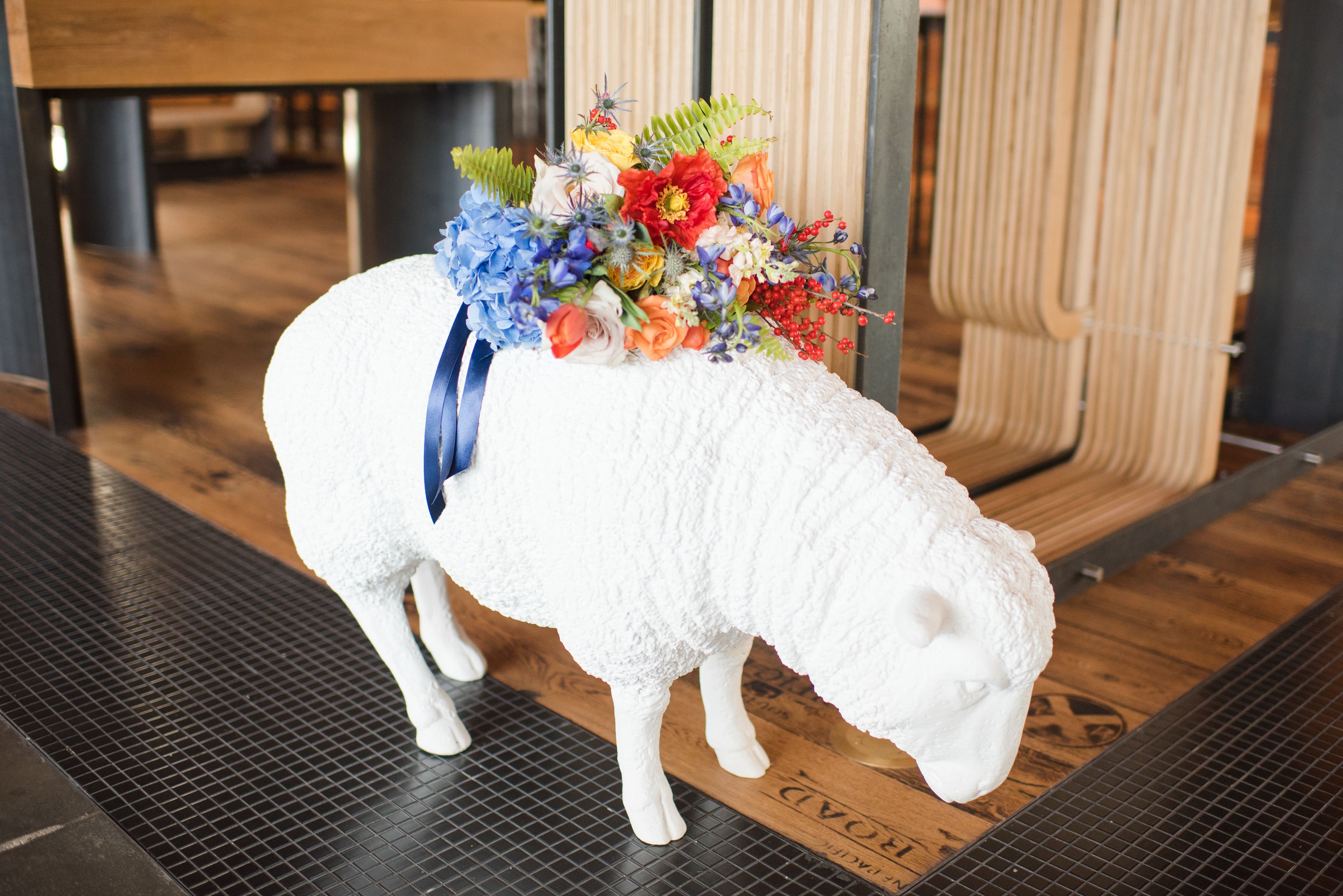 A white sheep sculpture with a colorful flower arrangement on its back, placed on a textured floor with wooden elements in the background.