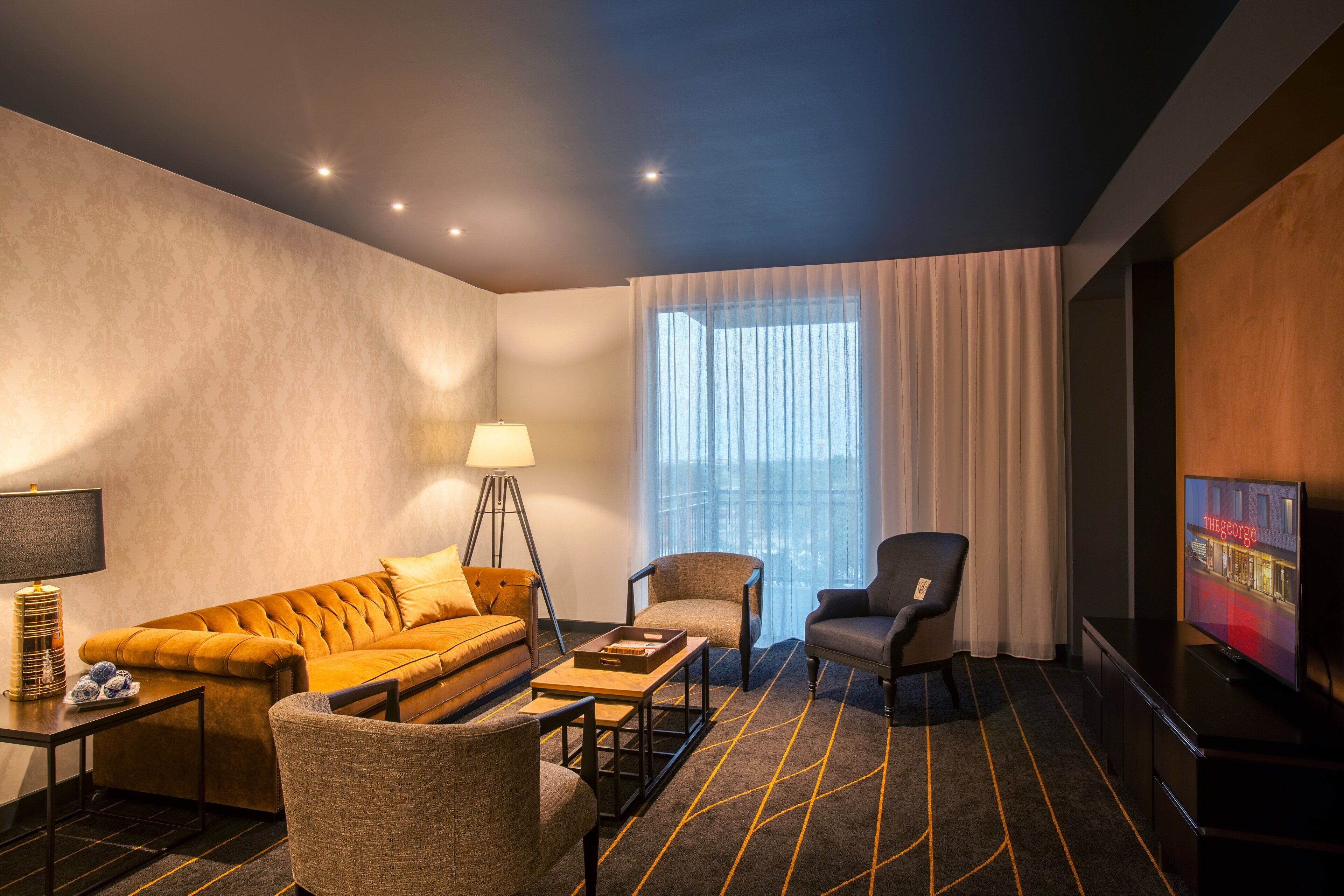 A hotel room with a sofa, armchairs, a coffee table, a lamp, a TV, and a large window with curtains.