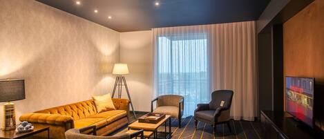 A hotel room with a sofa, armchairs, a coffee table, a lamp, a TV, and a large window with curtains.