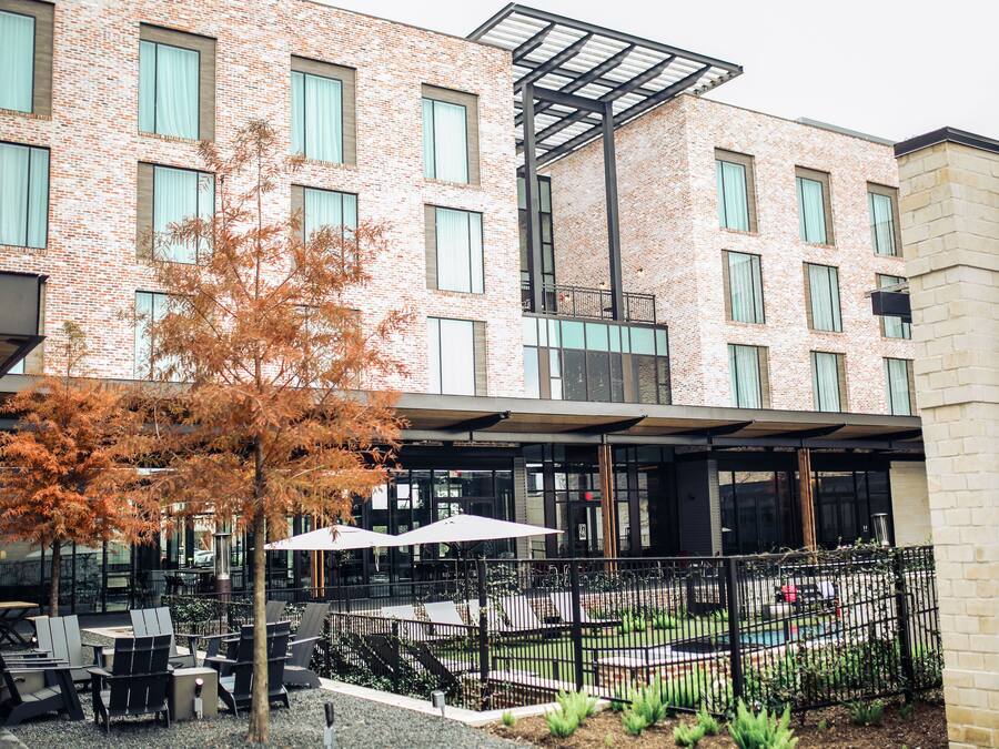 A modern building with a brick facade, outdoor seating area with tables and chairs, and a small pool.