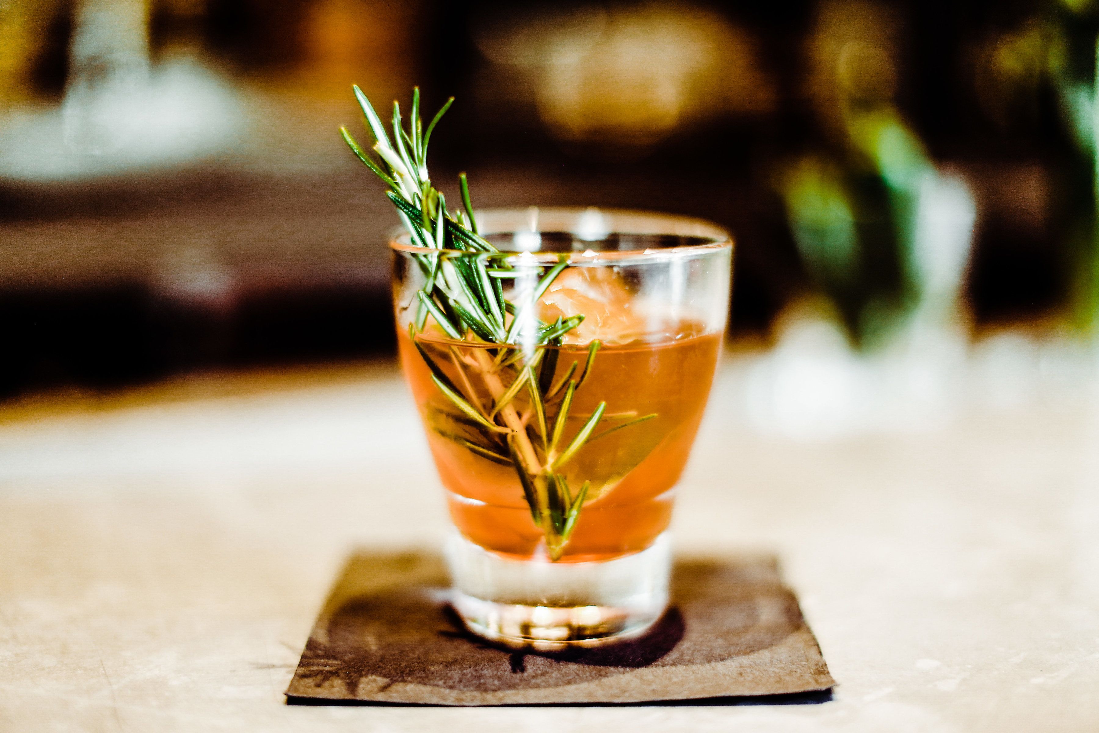 A glass of amber-colored beverage with a sprig of rosemary, placed on a coaster.