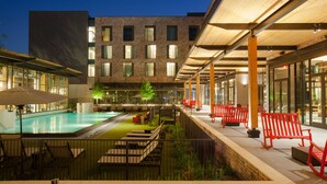 A modern outdoor pool area with a glass barrier, red seating, and a building with multiple windows in the background.