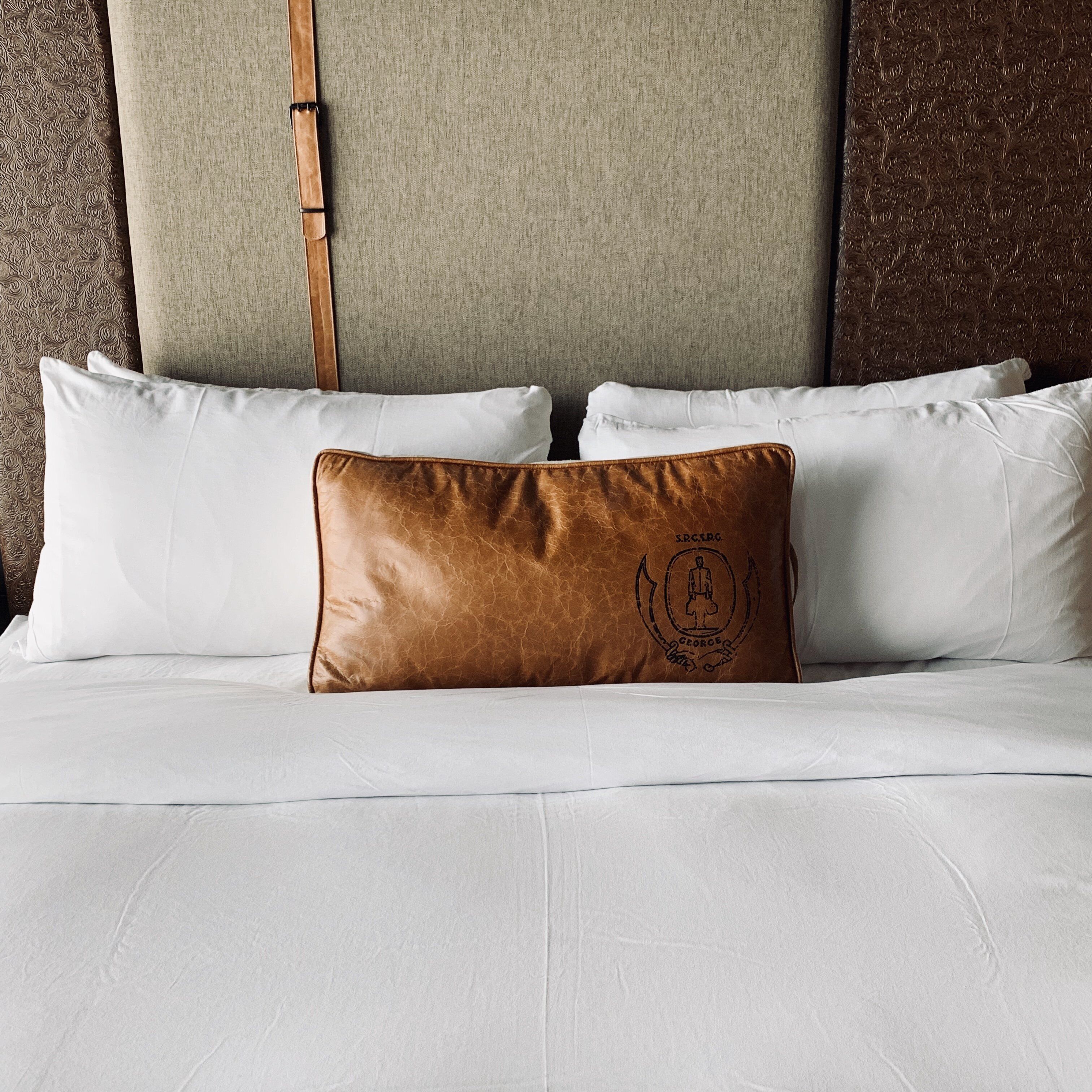 A bed with white bedding and a single brown pillow with a logo.
