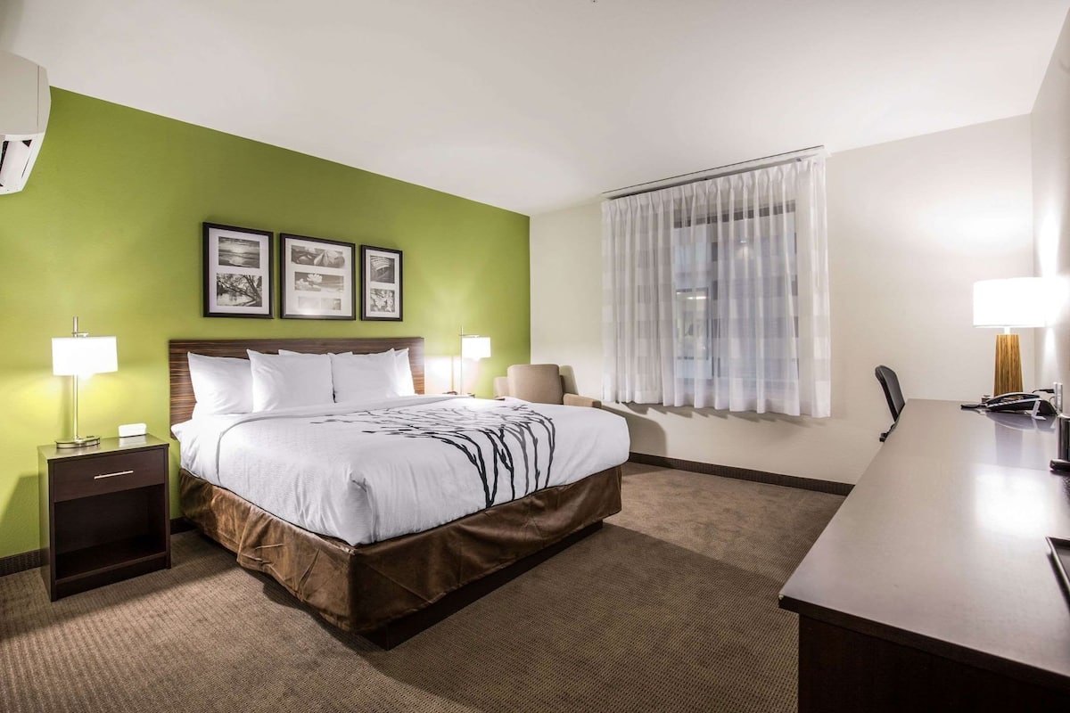 A hotel room with a bed, bedside tables, a desk, a chair, a window with curtains, and framed pictures on the wall.