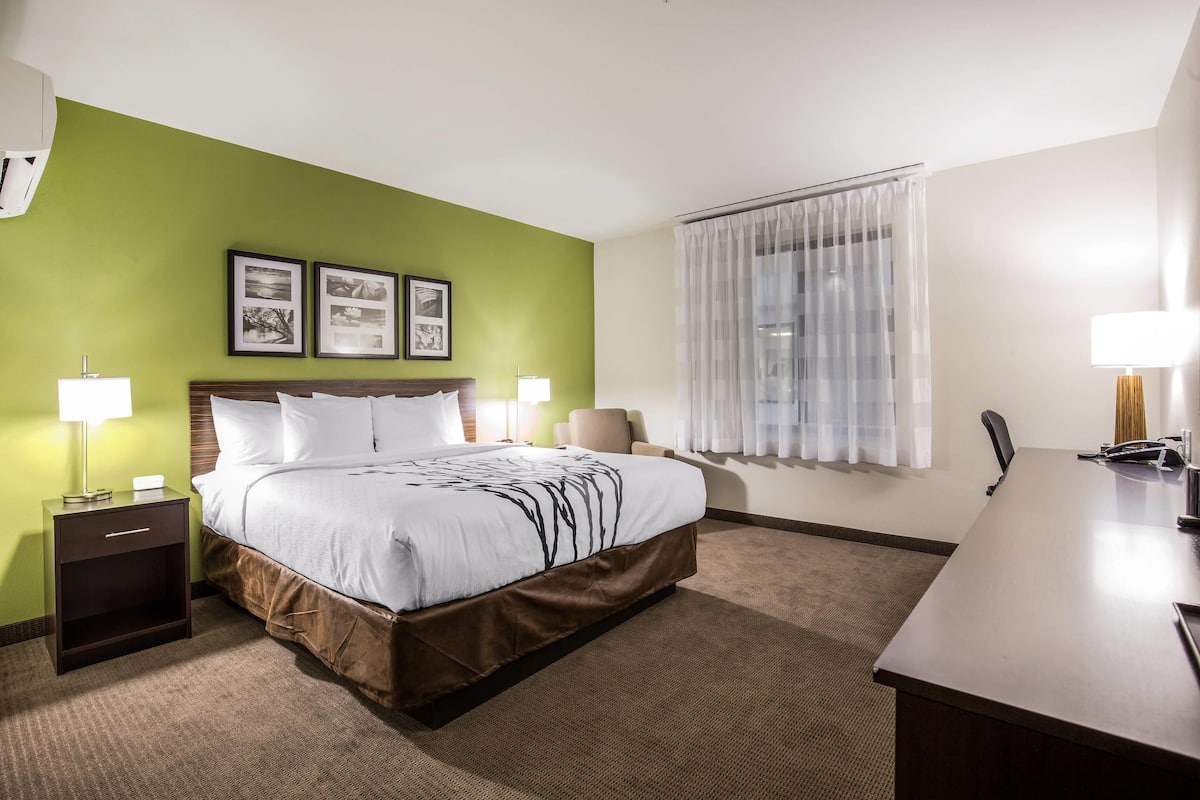 A hotel room with a bed, bedside tables, a desk, a chair, a window with curtains, and framed pictures on the wall.