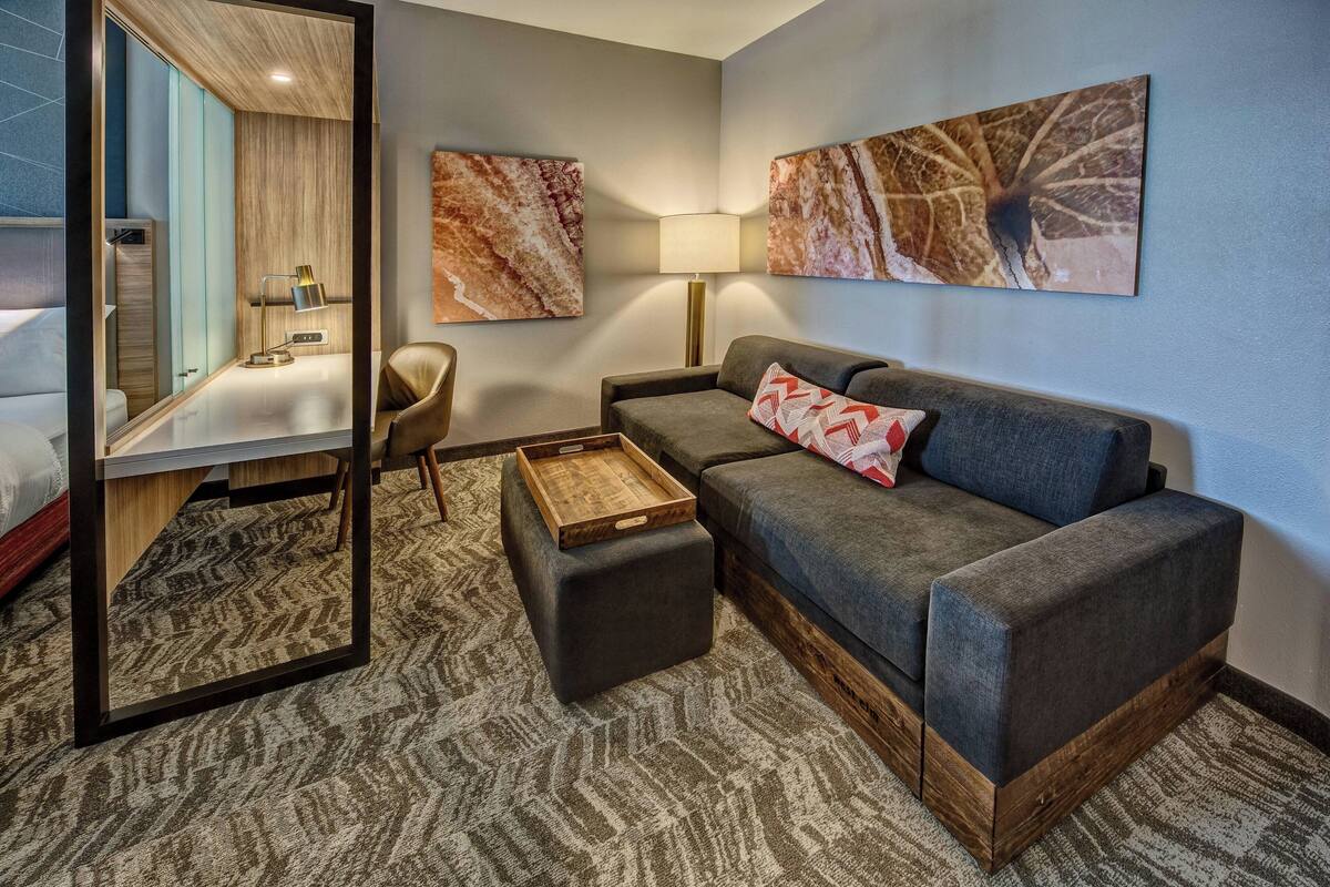 A modern hotel room with a sofa, a wooden side table, a chair, and a large mirror.
