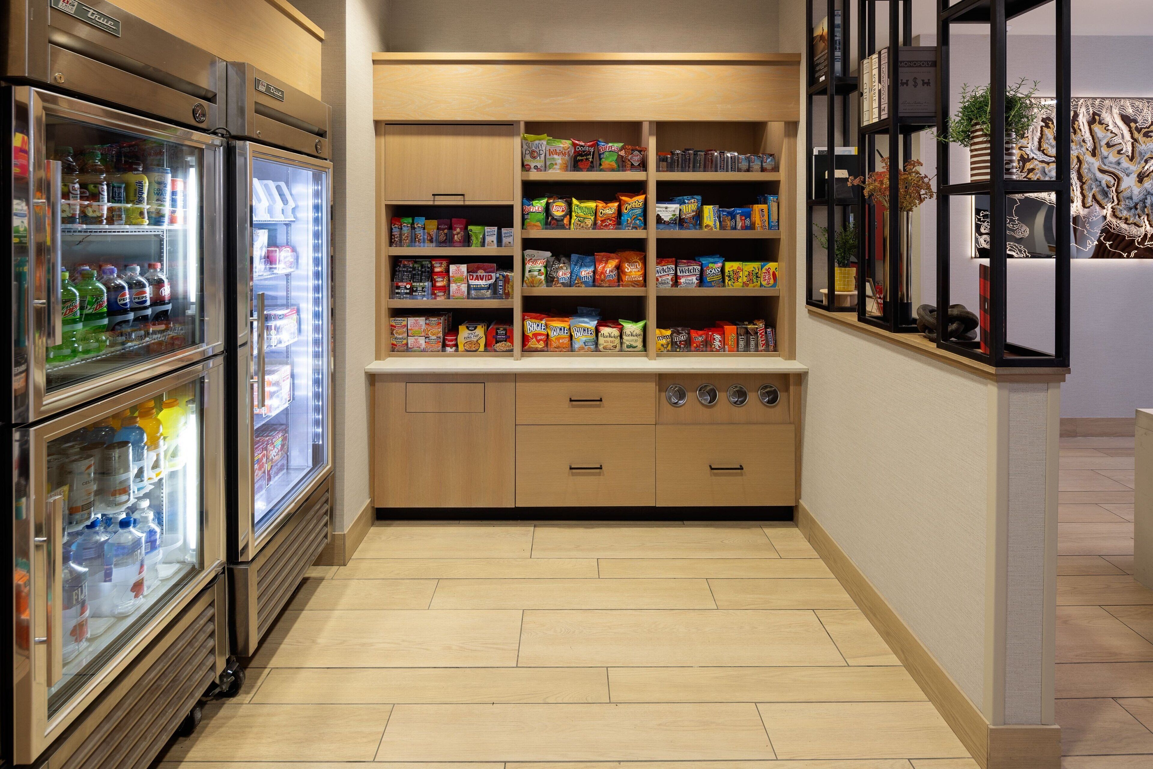 A well-lit corridor with a refrigerated beverage display, a snack shelf, and a decorative wall piece.