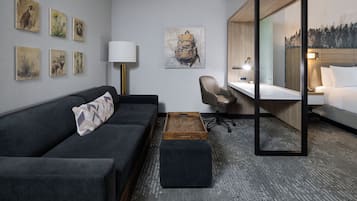 Quarto de hotel moderno com sofá cinza escuro, mesa lateral de madeira, escrivaninha com cadeira, espelho grande e obras de arte nas paredes.
