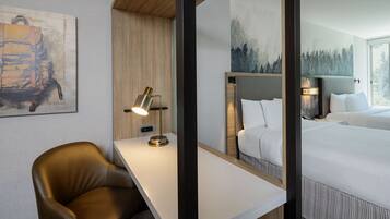 A hotel room with a wooden desk, a leather chair, a bed with white bedding, and a large mirror reflecting the room.