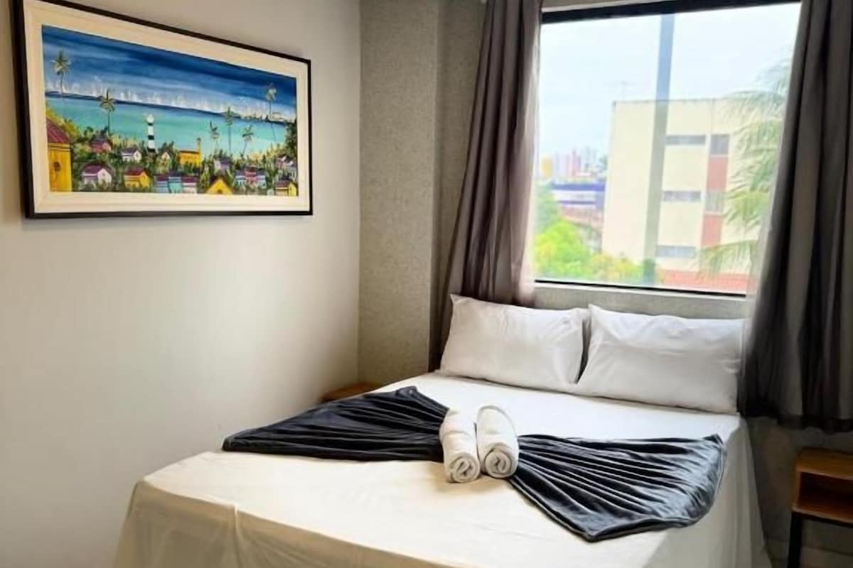 A hotel room with a bed, a painting on the wall, and a window with curtains.